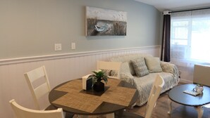 A living room with a round dining table, white chairs, a sofa with patterned cushions, and a painting on the wall.
