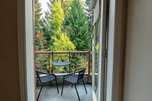 A balcony with a table and two chairs, a metal railing, and a view of trees and a building. - Alpenglow Lodge Large Studio by MVA (whistler)