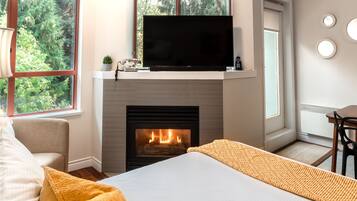 Una habitación con chimenea, un televisor en un estante, una cama, un ventanal con vistas a árboles y un comedor.
