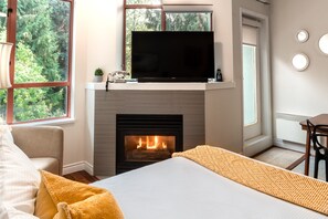 A room with a fireplace, a television on a shelf, a bed, a window with a view of trees, and a dining area. - Alpenglow Lodge Large Studio by MVA (whistler)