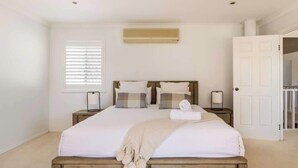 Un dormitorio con cama de madera, ropa de cama blanca, ventana con persianas, unidad de aire acondicionado y puerta que da a otra habitación.