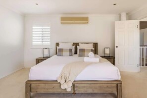 Un dormitorio con cama de madera, ropa de cama blanca, ventana con persianas, unidad de aire acondicionado y puerta que da a otra habitación.