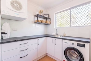 A compact kitchen with a built-in washing machine, a shelf with baskets, and a window with a mesh screen.