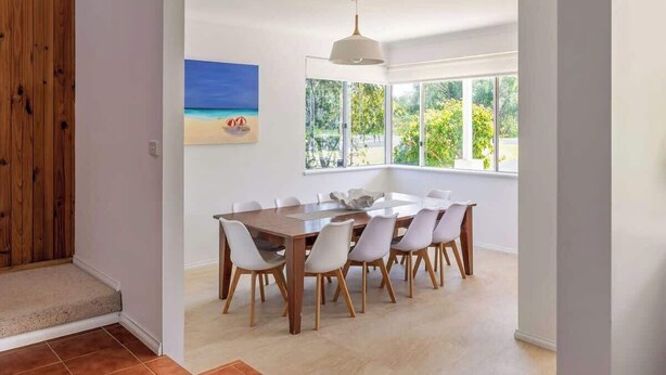 Un área de comedor con una mesa grande y sillas, un cuadro de una playa y ventanas amplias con vista a follaje.