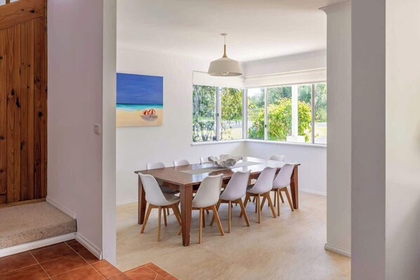 Un área de comedor con una mesa grande y sillas, un cuadro de una playa y ventanas amplias con vista a follaje.