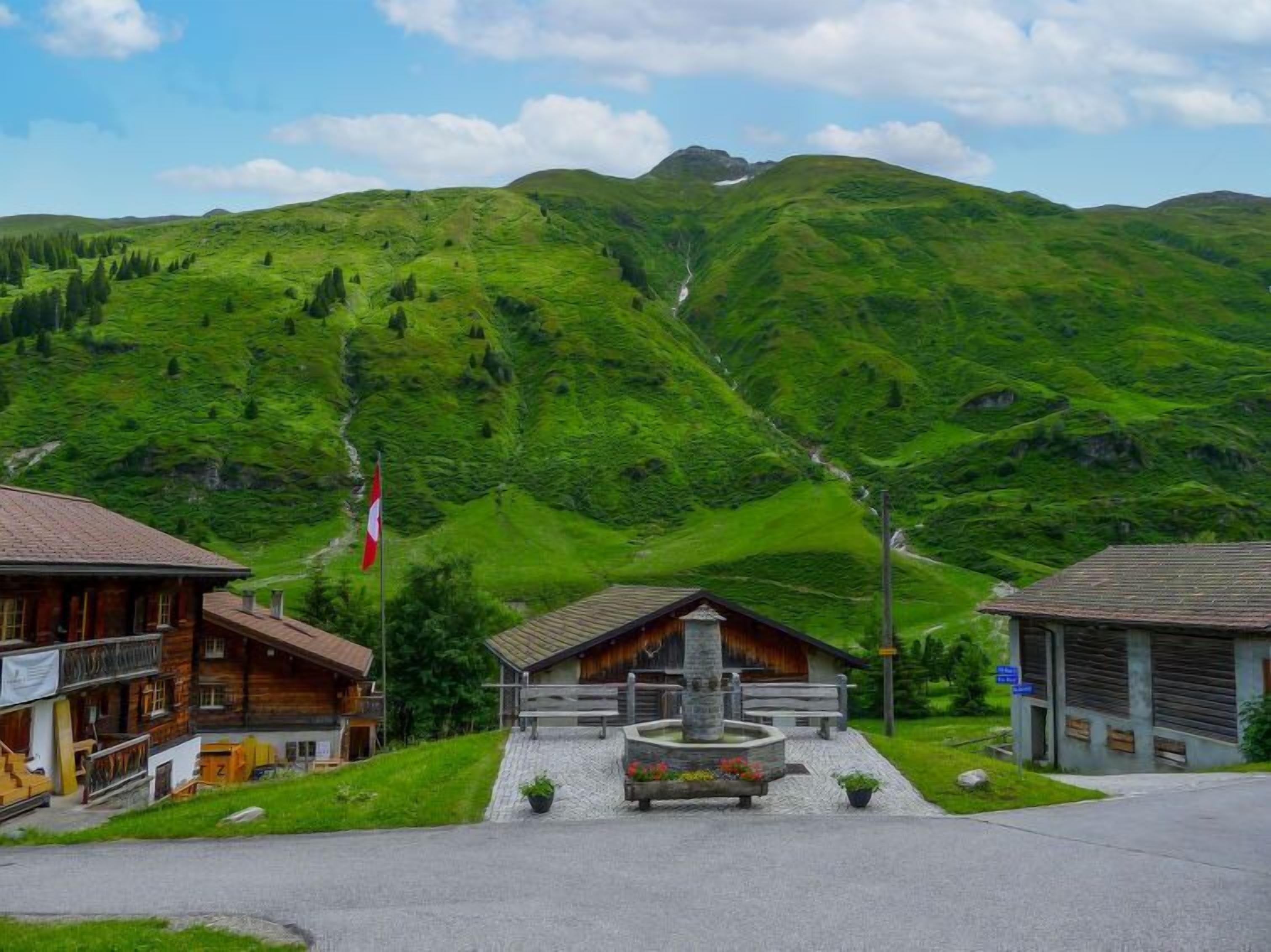A mountain village with wooden buildings, a flag, and a stone fountain.