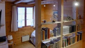Ein gemütliches Blockhaus-Interieur mit Holzwänden, einer Bücherwand voller Bücher, einem Bett und einem Fenster mit Vorhängen.