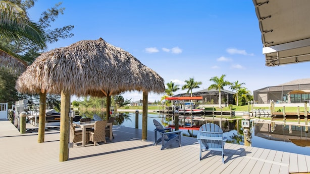 A wooden deck with a thatched umbrella, outdoor seating, and a view of a marina with boats.