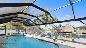 An indoor swimming pool with a clear view of the surrounding area, including palm trees and a thatched-roof structure.