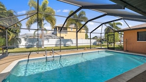 Une piscine d’un bleu éclatant, protégée par une paroi de verre, entourée de palmiers et d’une clôture blanche.