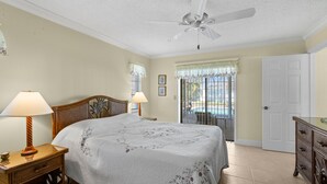 Une chambre à coucher avec un lit, deux lampes de chevet, un ventilateur de plafond, une vue sur une piscine par la fenêtre et une porte.