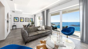 Ein modernes Wohnzimmer mit einem grauen Sofa, einem Glastisch und einem blauen Sessel. Ein großes Fenster bietet Blick auf das Meer.