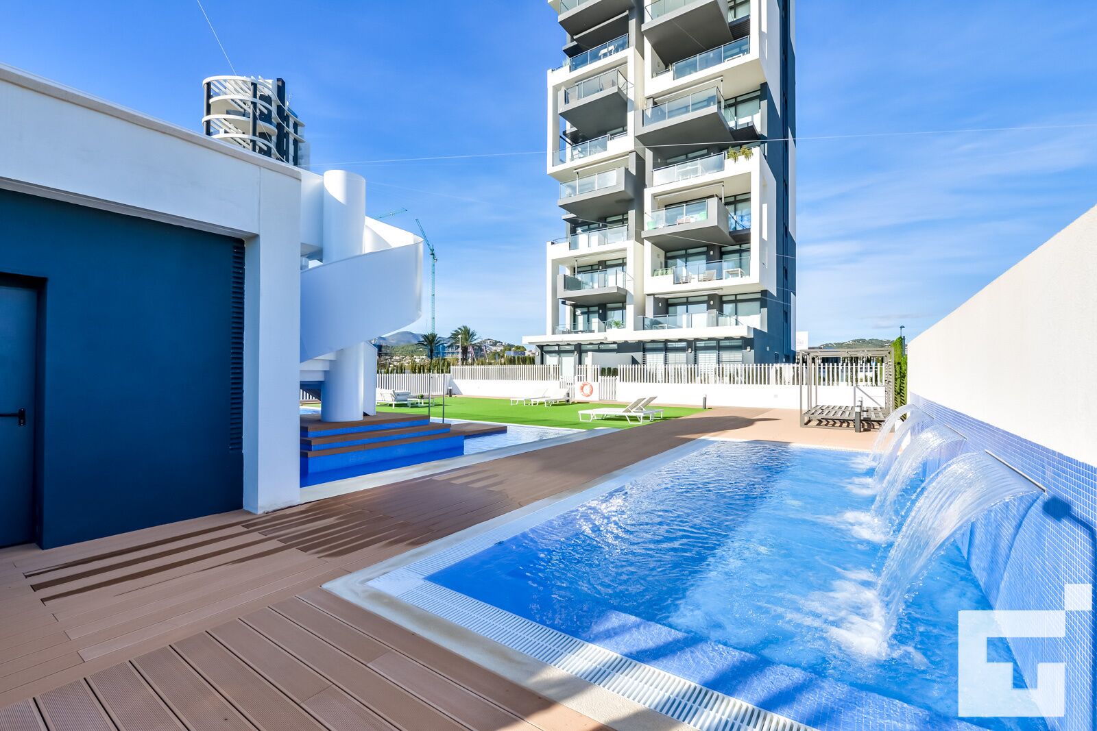 Un hotel moderno con piscina, un área exterior techada y un edificio de varios pisos al fondo.