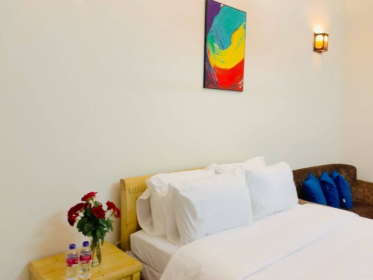 A hotel room with a bed, white bedding, a nightstand with a vase of red flowers, and a colorful abstract painting on the wall.