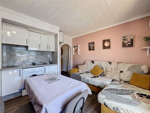 A compact living space with a kitchenette, a sofa with patterned cushions, and a dining table with a metal charger. - JARDINS II 1-9 Roses / Roses - INTERimmo (Rosas / Roses)