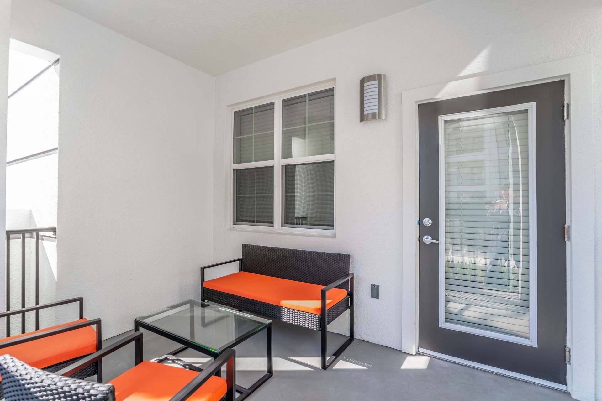 A balcony with a glass table and two chairs, a dark door with a window, and a white wall with a window.