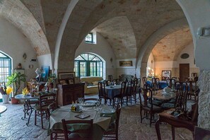 Un espace repas comprenant plusieurs tables et chaises, des arches en pierre et de grandes fenêtres laissant entrer la lumière naturelle.