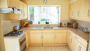 A kitchen with wooden cabinets, a gas stove, a sink, and a window with a view of greenery. - Beautiful 3 Bedroom Home in Staines Private Parking, Garden, Near Heathrow (Staines-upon-Thames)