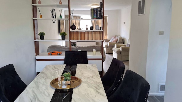 A dining area with a marble table, black chairs, and a tray with a plant and candles. There are shelves with books and decorative items in the background. - Beautiful 3 Bedroom Home in Staines Private Parking, Garden, Near Heathrow (Staines-upon-Thames)