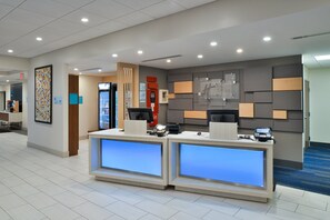 Ein Hotel-Empfangsbereich mit zwei Rezeptionen, einem Feuerlöscher, einem Automaten und einer Wandtafel mit Informationen.