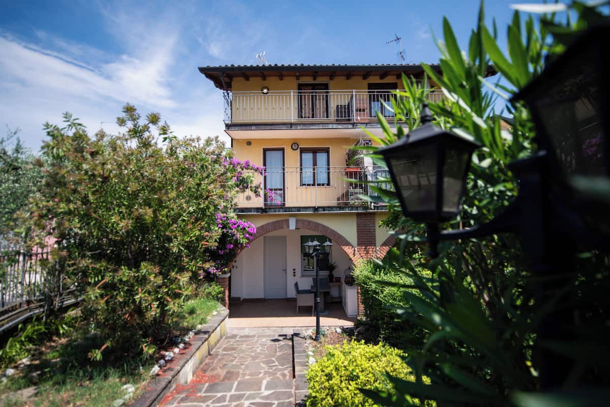 Un bâtiment à deux étages doté d’un balcon, d’un chemin de pierre menant à l’entrée et entouré de verdure.