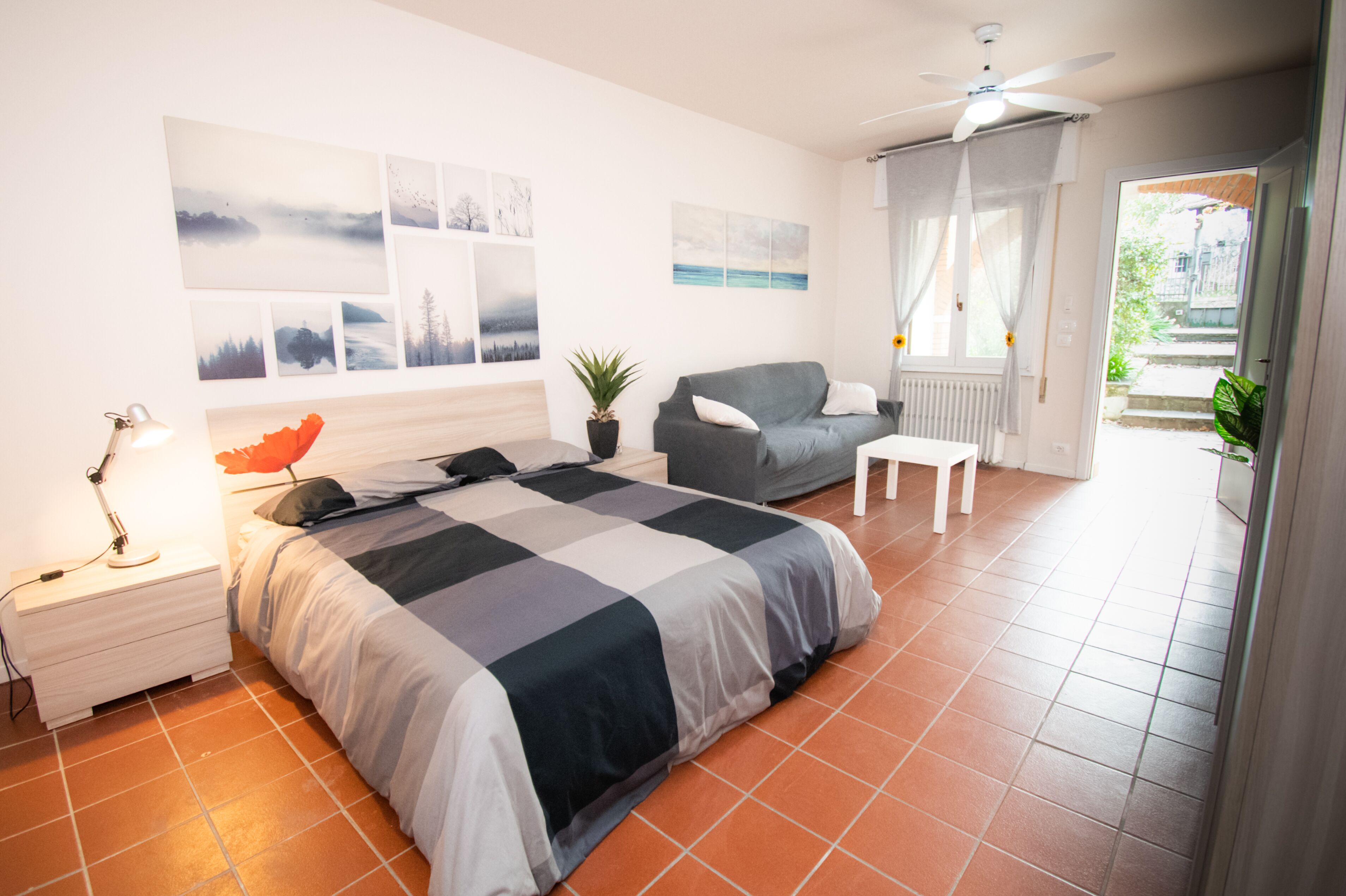 Une chambre avec un grand lit, un fauteuil gris, une petite table, une table de chevet avec une lampe et un ventilateur de plafond.