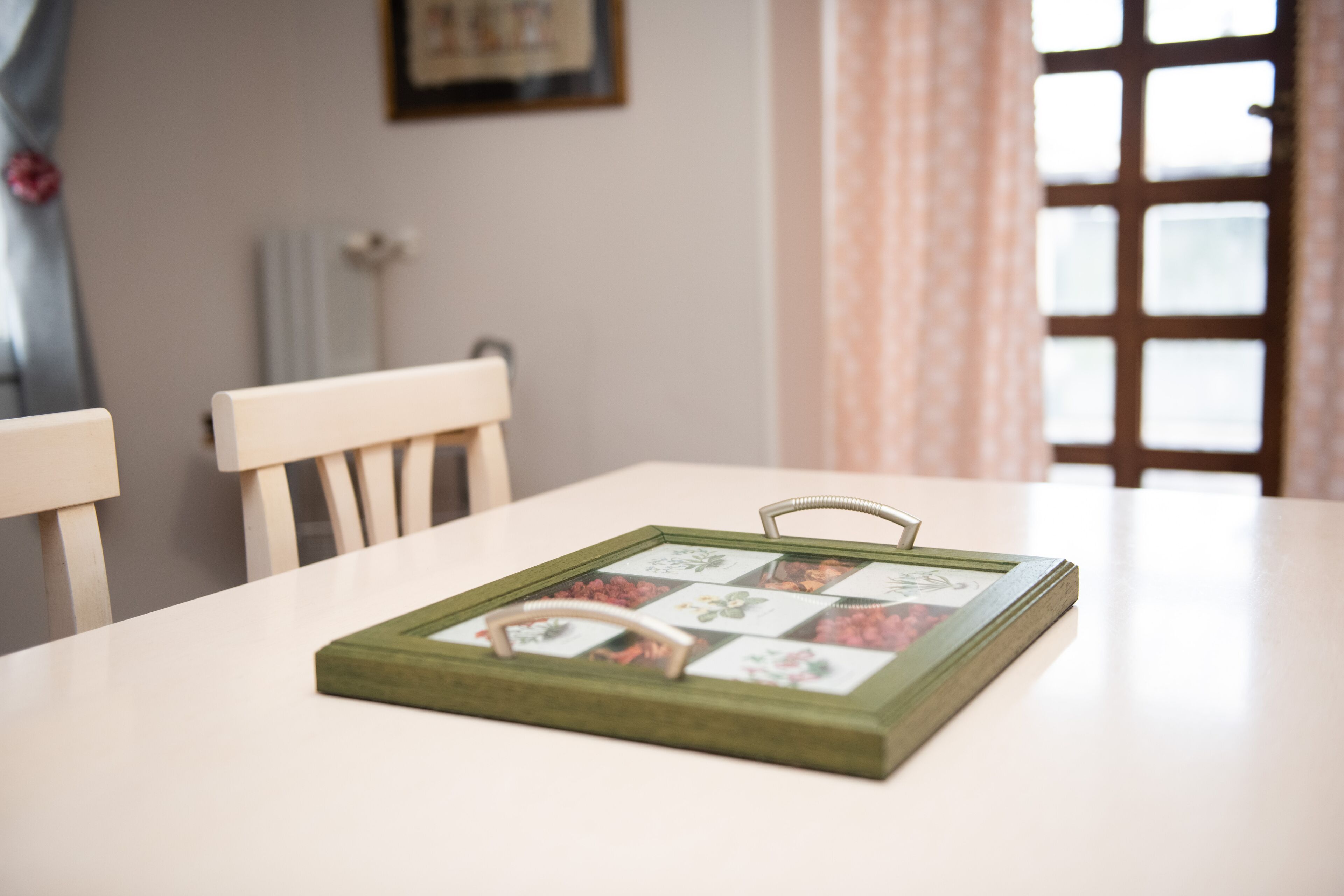A framed collage on a table with a handle, a white chair, and a window with curtains.