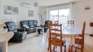 Ein Wohnzimmer mit einer schwarzen Ledersofa, einem Glastisch und einem Esstisch mit Stühlen. Zwei Pendelleuchten, ein großes Fenster und Wandbilder sind vorhanden.