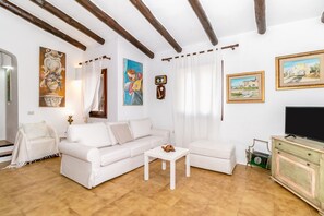 A living room with a white sofa, a white coffee table, and a television on a cabinet. There are paintings on the walls and a window with curtains.