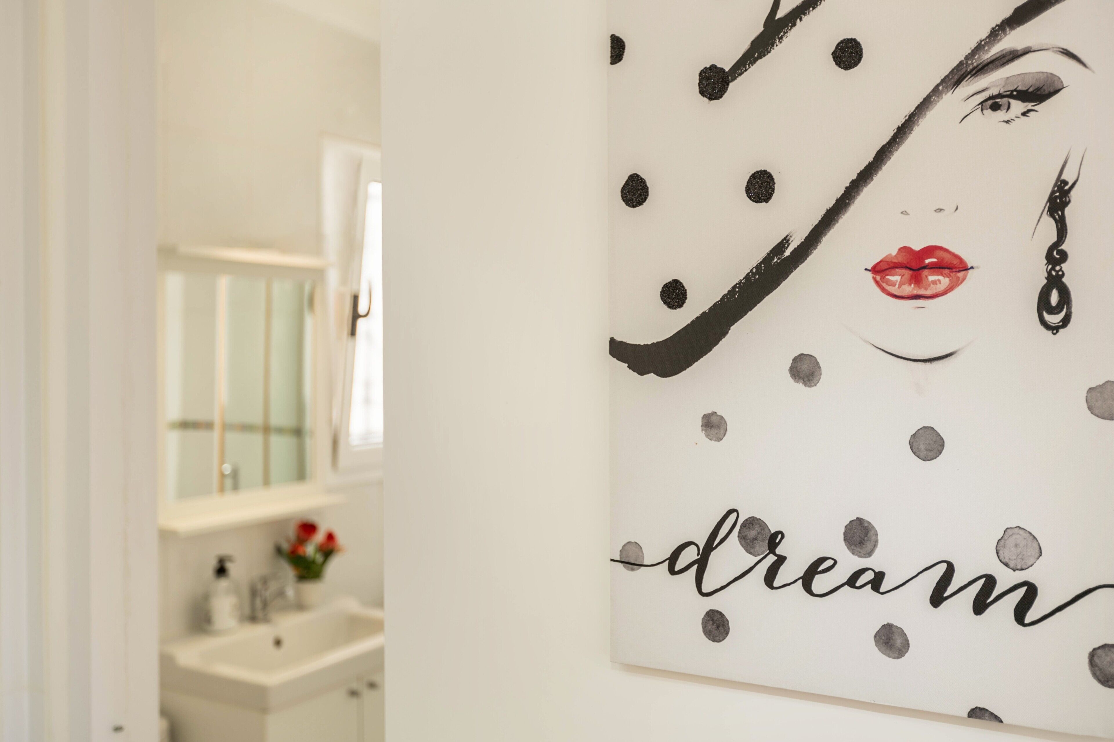 Une salle de bain avec un lavabo blanc, un miroir et une œuvre d’art murale représentant le visage d’une femme, avec le mot « rêve » écrit en dessous.