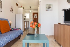 Una sala de estar con un sofá, una mesa de centro con un florero, un televisor y una cocina al fondo.