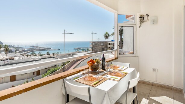 Un balcon avec une table dressée pour le dîner, offrant une vue sur la côte, avec des bâtiments et une jetée.