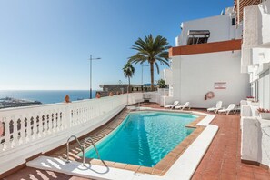 Una piscina en la azotea con vista al mar, paredes blancas y una palmera.