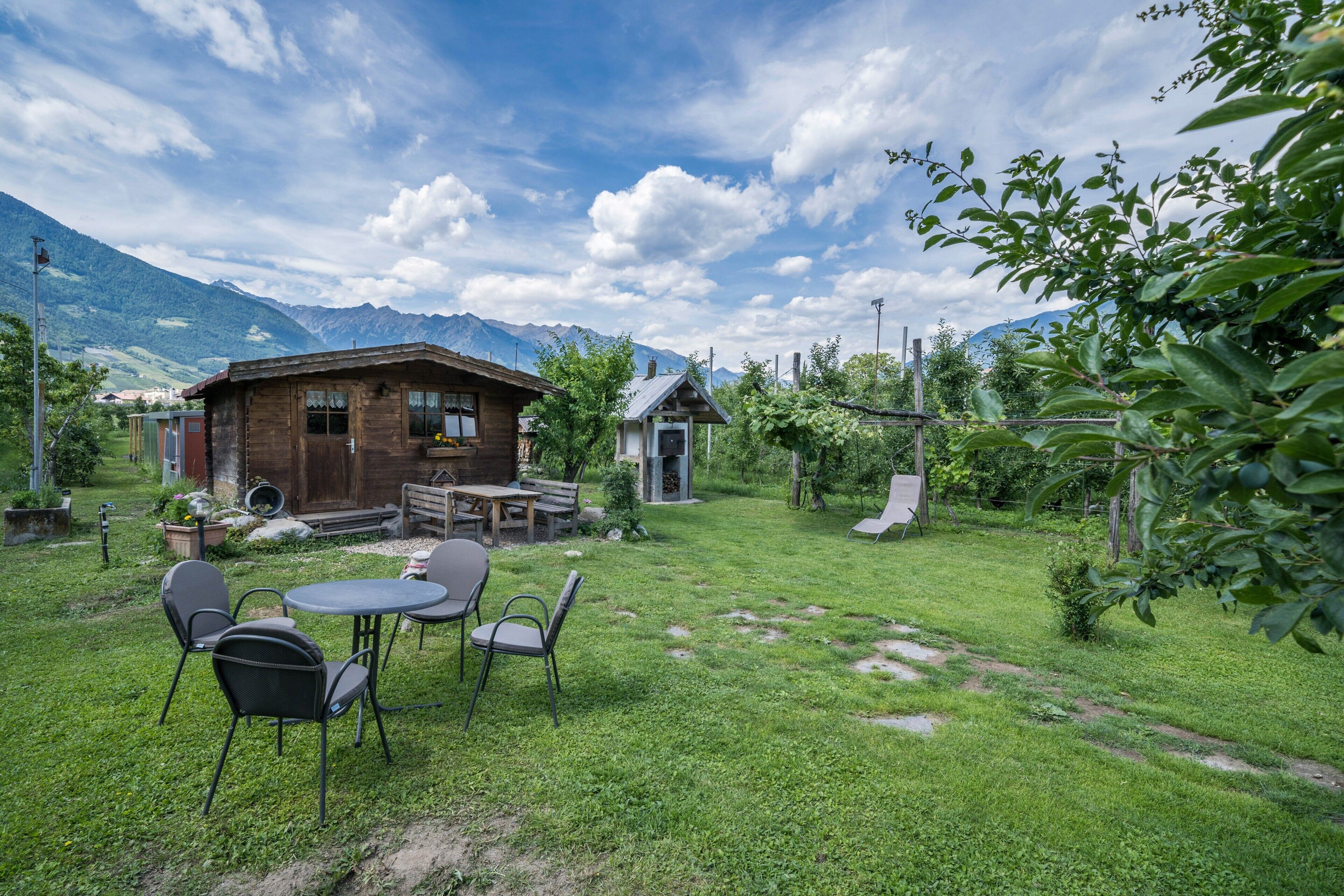 Una casa de madera rústica con un patio, rodeada de abundante vegetación y montañas al fondo.