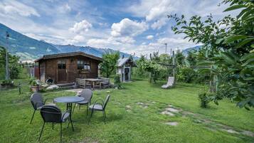 Una casa de madera rústica con un patio, rodeada de abundante vegetación y montañas al fondo.