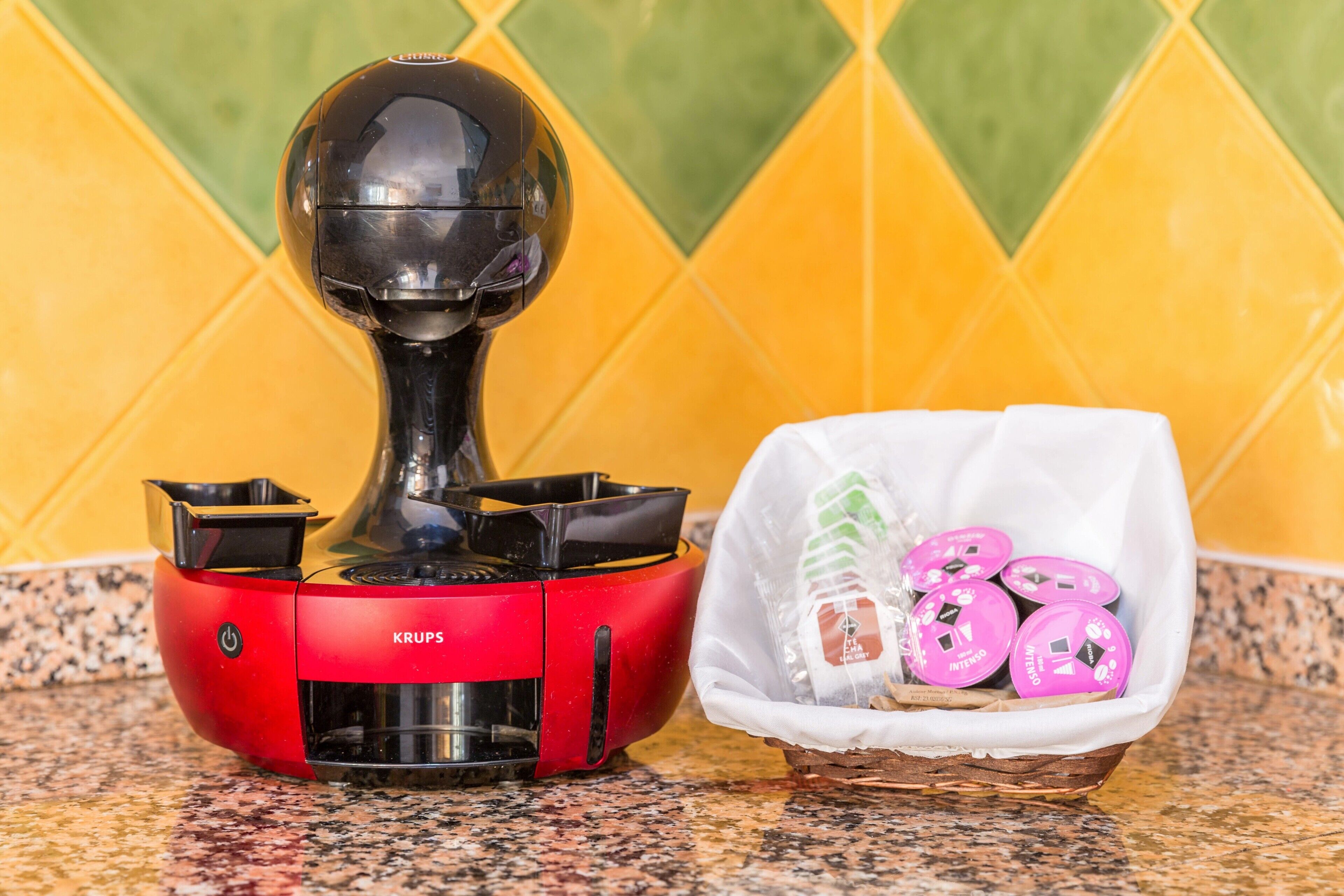 A red Krups electric kettle with a transparent lid, a white plastic bag containing pink containers, and a yellow and green tiled backsplash.