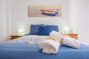 A neatly made bed with blue bedding, a painting of a boat on a beach, and two bedside tables with lamps. - Cozy Apartment "Las Playitas" near the Beach with Wi-Fi & Terrace (Tuineje)