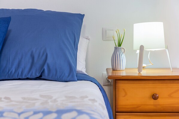 A bed with blue bedding, a wooden nightstand with a vase of flowers, and a white lamp. - Cozy Apartment "Las Playitas" near the Beach with Wi-Fi & Terrace (Tuineje)