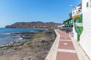 A coastal walkway with a rocky beach, a mountain in the background, and buildings with balconies. - Cozy Apartment "Las Playitas" near the Beach with Wi-Fi & Terrace (Tuineje)