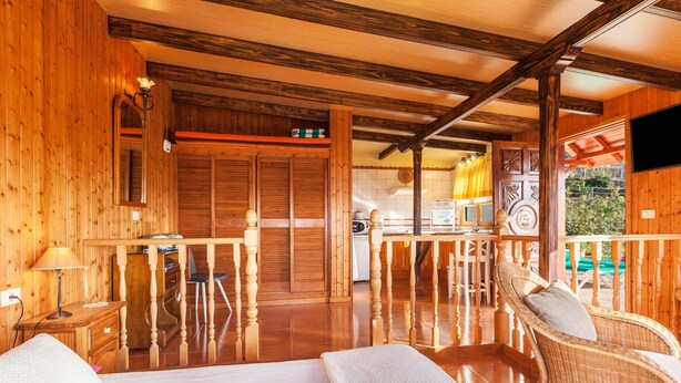A wooden cabin interior with a bed, a dining area, and a kitchen visible through an open door.