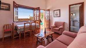 Uma sala de estar com sofá, mesa de jantar com cadeiras, mesa de centro e janela com porta aberta para o exterior.
