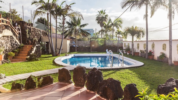 Piscina rodeada por palmeiras e um muro de pedra, com uma escadaria que leva a um edifício.