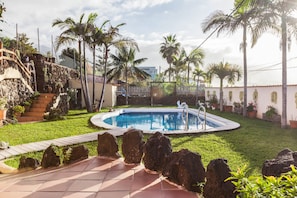 Una piscina rodeada de palmeras y un muro de piedra, con una escalera que sube hacia un edificio.