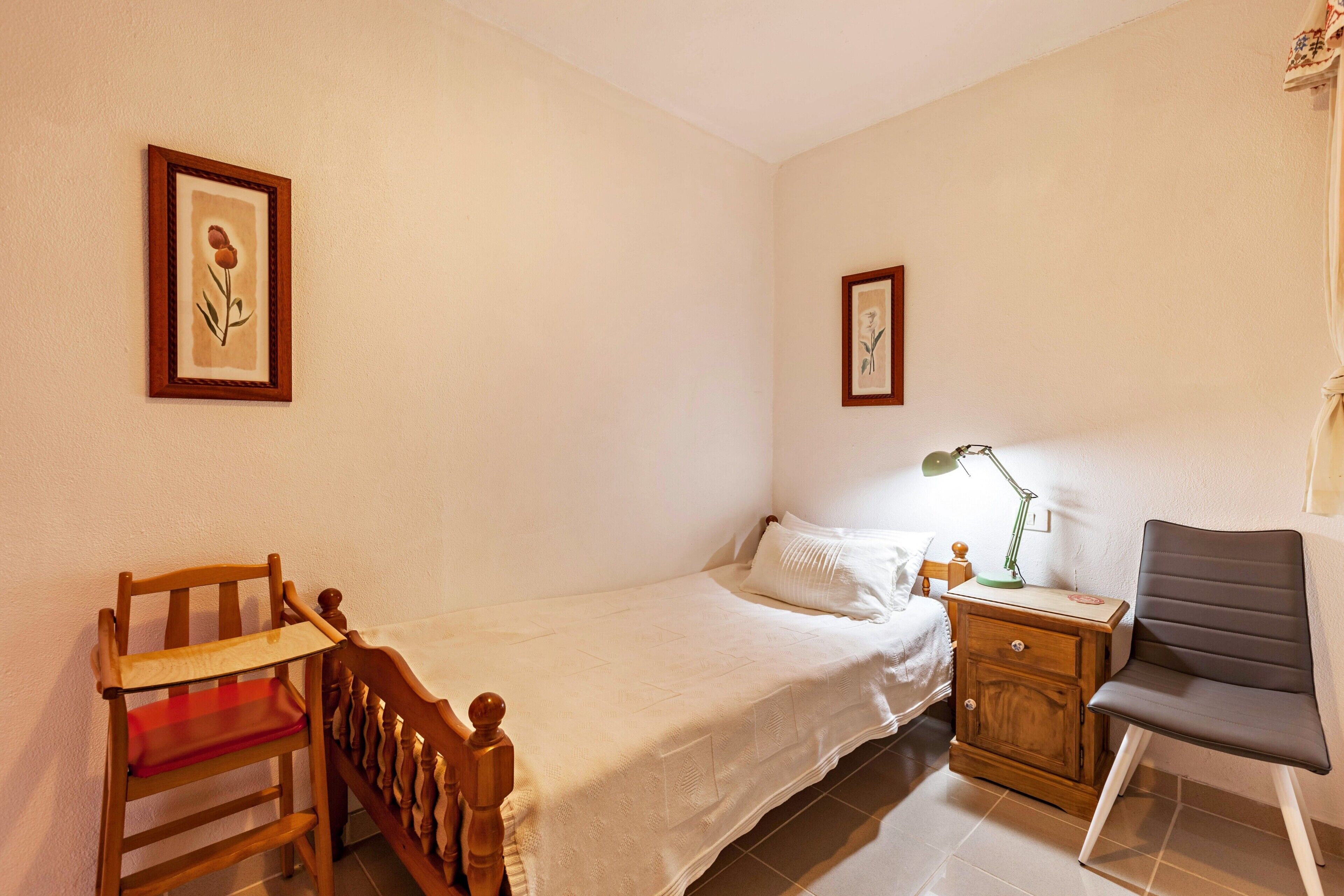 Una cama individual con cubrecama blanco, una mesita de noche de madera con lámpara, una silla y dos cuadros en la pared.