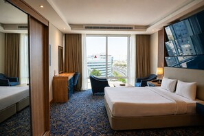 A hotel room with a large bed, a desk, a chair, and a view of the cityscape through large windows. - Almansour Suites Hotel Doha (Doha)