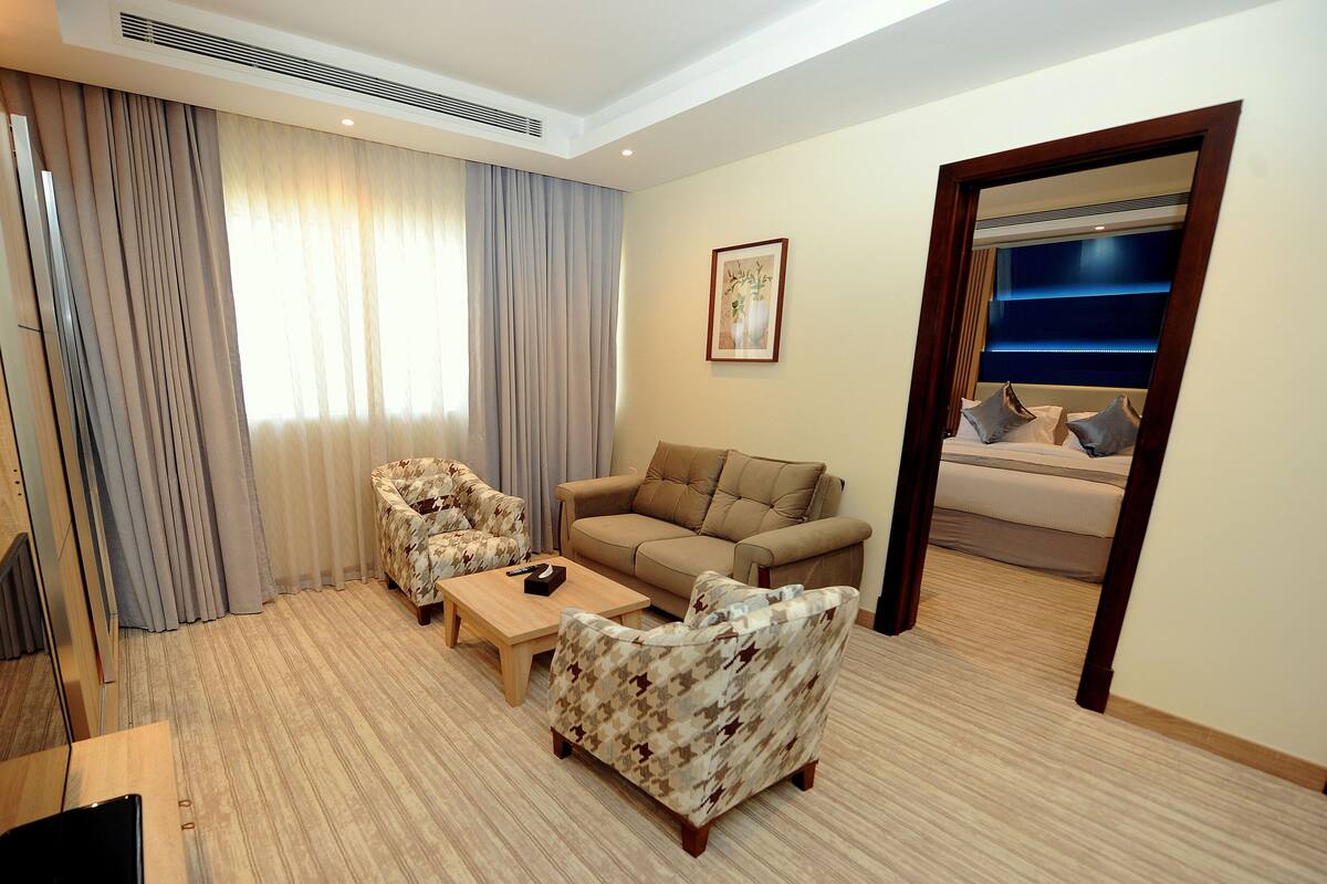 A hotel room with a sofa, armchair, and a small table. There is a bed with a headboard and a painting on the wall.