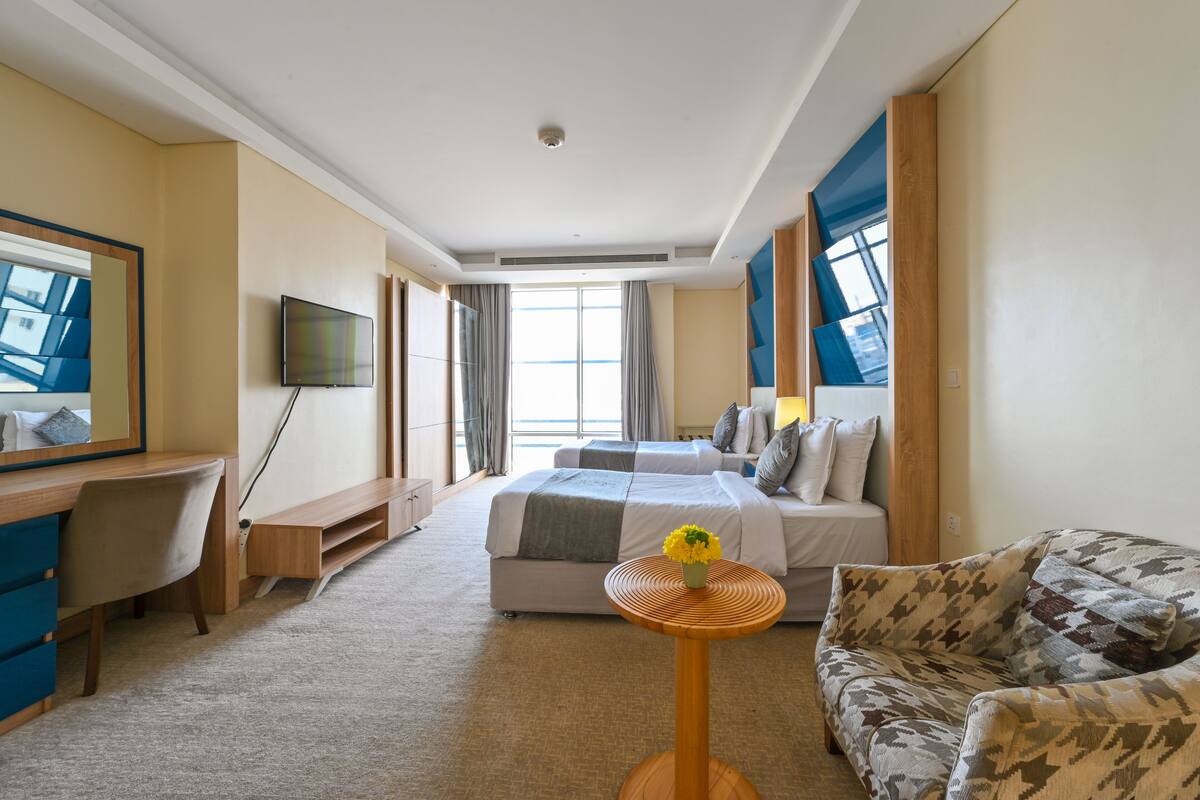 A hotel room with a bed, a desk, a chair, a sofa, a TV, and a window with curtains.
