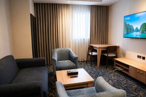 Une chambre d’hôtel avec un canapé, un fauteuil, une petite table avec un livre, un téléviseur fixé au mur et une fenêtre avec des rideaux.
