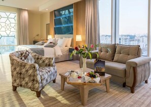 Habitación de hotel con cama, sofá, sillón, una mesita de centro con frutas y flores, y vistas a la ciudad a través de amplios ventanales.