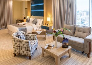 Ein Hotelzimmer mit Bett, Sofa, Sessel, Nachttisch, Lampe, Blumen in einer Vase und einer Zeitschrift auf einem Tisch.